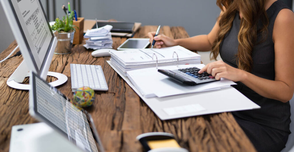Person using a calculator while reviewing documents and financial reports at a desk.