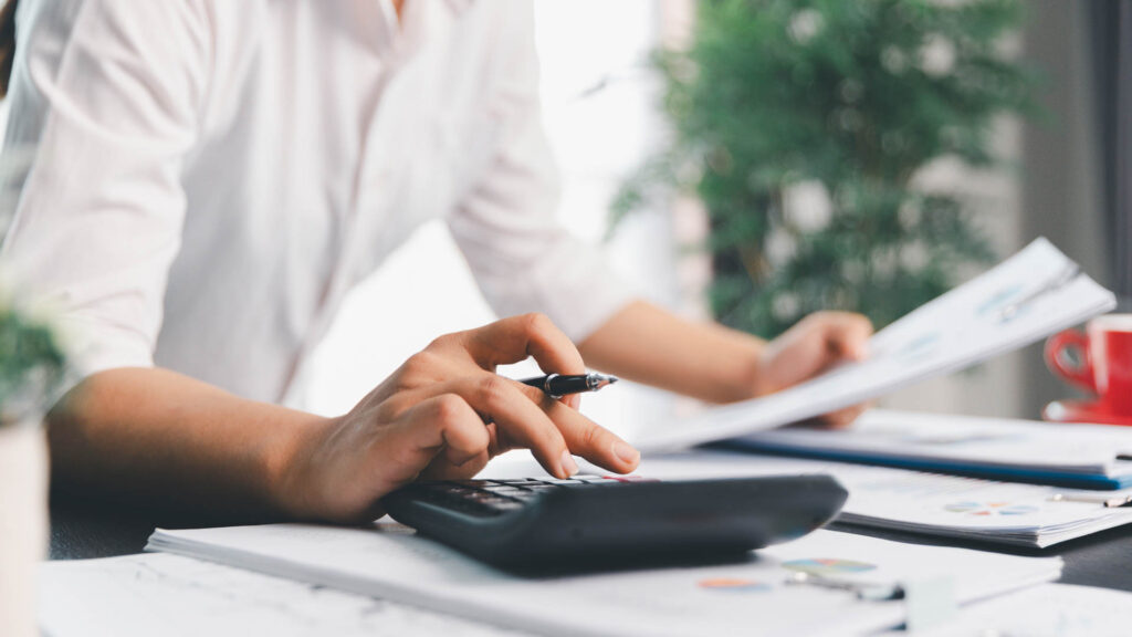 Person calculating figures on a calculator next to a laptop and financial documents.