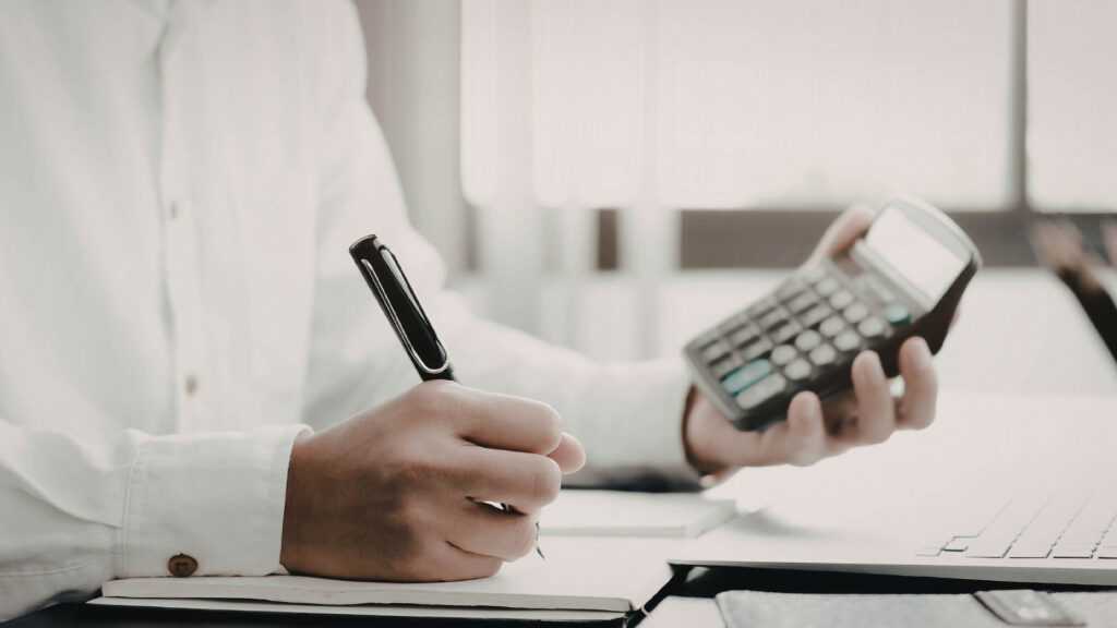 Person calculating figures on a calculator next to a laptop and financial documents.
