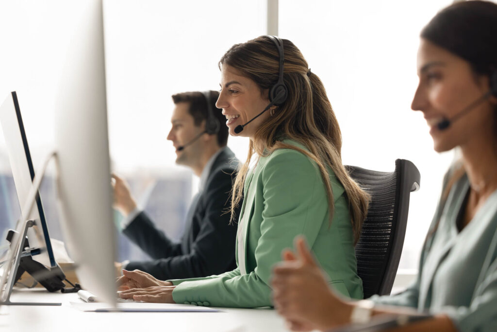 Group of customer service representatives sit at modern office workstation, focus on young Hispanic woman wearing headset engaged in conversation, assisting client or participating in virtual meeting