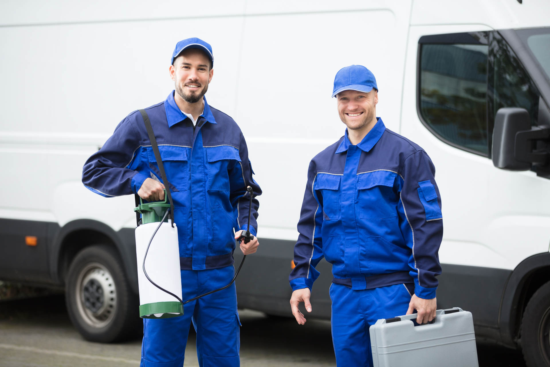 Two pest control technicians in blue uniforms standing by a service van with equipment.