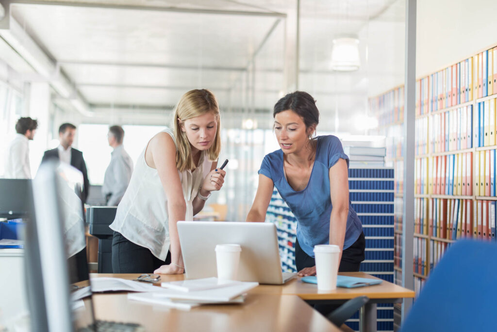 Assistant and partner coffee meeting. Focus on two women in her 30s and 40s who are standing in front of a laptop and two white coffee cup, in a luminous open space looking at balance sheets